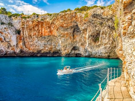 Türkisfarbenes Wasser an der Grotta Zinzulusa in Castro mit Kalksteinklippen, einem Boot in der Bucht und Steinstufen mit Metallgeländer.