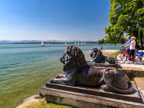 Zwei Radfahrer mit Rädern neben bronzenen Löwenstatuen am Starnberger See. Segelboote auf dem See, Berge im Hintergrund unter blauem Himmel.