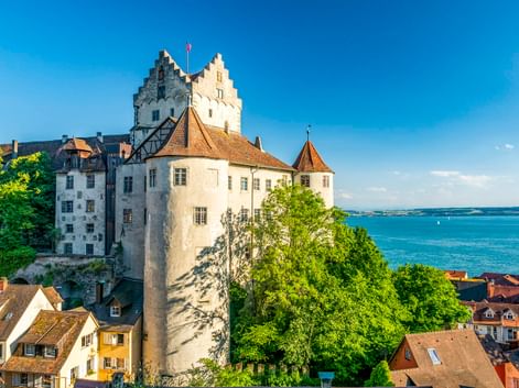 Historische Burg Meersburg mit weißen Mauern und rundem Turm mit Blick auf den Bodensee. Häuser mit roten Dächern im Vordergrund, grüne Bäume.