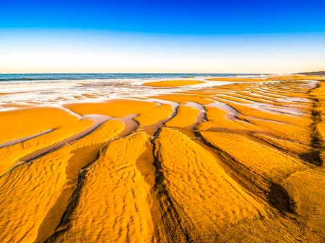 Breiter Sandstrand in Mimizan Plage mit gewellten goldenen Sandmustern und flachen Wasserpfützen. Der Atlantik trifft auf klaren blauen Himmel.