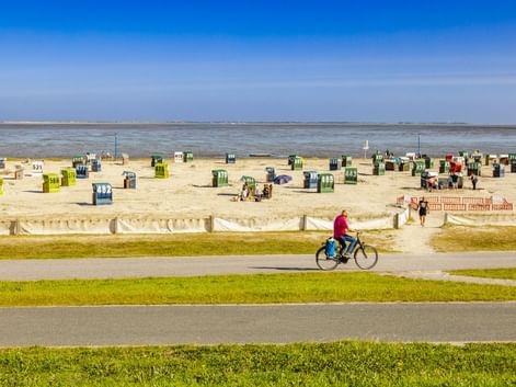 Radfahrer auf Weg entlang Neuharlingersiel Strand mit bunten Strandkörben im Sand, Nordseeküste im Hintergrund unter blauem Himmel.