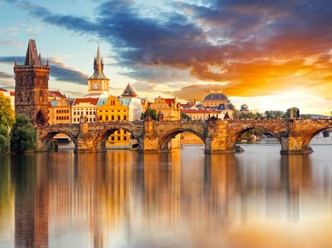 Karlsbrücke über die Moldau in Prag bei Sonnenuntergang mit historischen Türmen und Gebäuden, die sich im ruhigen Wasser unter dramatischem Himmel spiegeln.