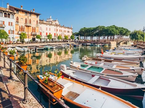 Hafen in Desenzano mit vertäuten Booten entlang der Promenade. Historische Gebäude säumen das Ufer, Bäume und eine Brücke im Hintergrund.