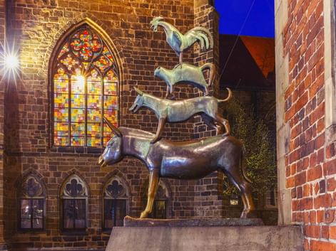 Bronze statue of Bremen Town Musicians showing stacked animals (donkey, dog, cat, rooster) illuminated at night beside a Gothic church with stained glass.