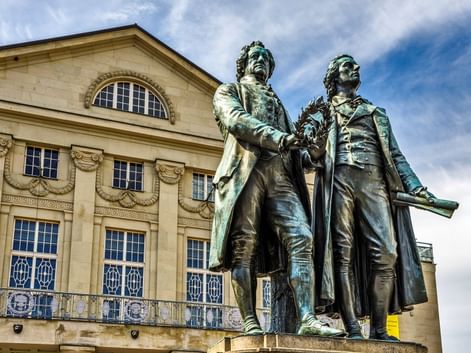Bronzestatue zweier Männer in Kleidung des 18. Jahrhunderts auf einem Sockel, vor einem neoklassizistischen Gebäude mit Bogenfenstern.