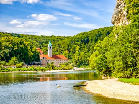 Weltenburg Abbey with bell tower beside the Danube River, surrounded by green forested hills. Kayakers paddle near a sandy beach in the foreground.