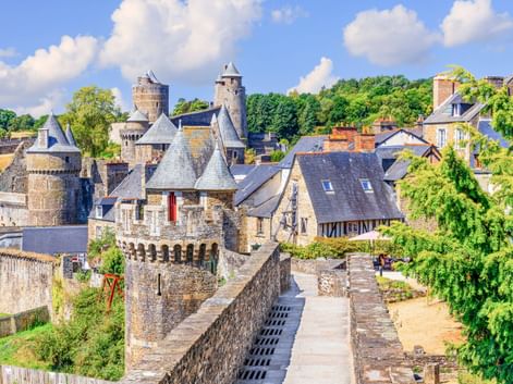 Burg Fougères mit Steintürmen und befestigten Mauern, umgeben von historischen Gebäuden und grüner Vegetation unter blauem Himmel mit weißen Wolken.