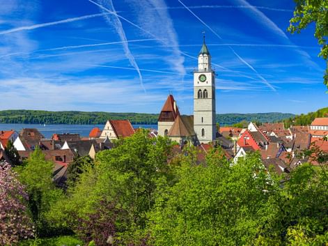 Panoramablick auf Überlingen mit weißem Kirchturm, roten Ziegeldächern und dem Bodensee im Hintergrund unter blauem Himmel.