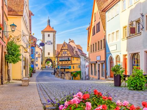 Cobblestone street in Rothenburg with colorful half-timbered houses and Siebersturm tower with orange roof. Pink and red flowers bloom in foreground.