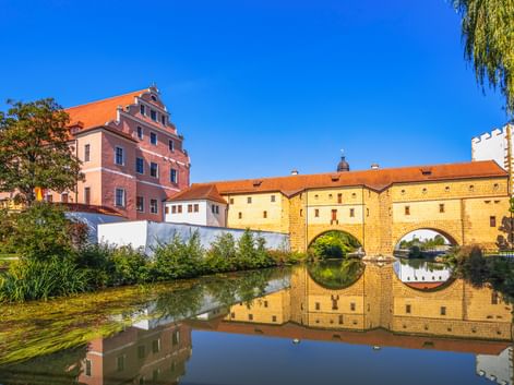 Historic Amberg Water Gate with double arches reflected in calm water. Pink Renaissance building and orange-roofed structures under blue sky.