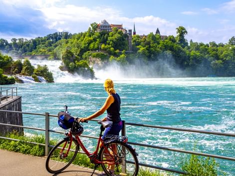 Radfahrerin mit rotem Fahrrad betrachtet den Rheinfall von einer Aussichtsplattform. Türkisfarbenes Wasser fließt an grünen bewaldeten Hügeln mit Schloss vorbei.