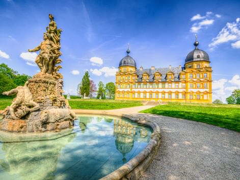 Gelbes barockes Schloss Seehof mit Kuppeltürmen hinter ornamentaler Brunnenskulptur. Kiesweg führt durch grüne Rasenflächen unter blauem Himmel.
