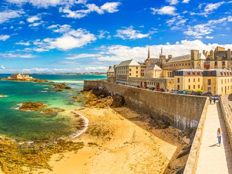 Sandstrand entlang historischer Steinmauern in Saint-Malo mit türkisfarbenem Wasser, Felsen und historischen Gebäuden unter blauem Himmel.