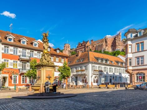 Kornmarkt in Heidelberg mit historischen Gebäuden, verzierter Brunnensäule und Blick auf die Schlossruine am Hang unter blauem Himmel.