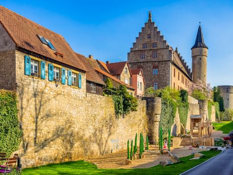 Medieval town wall in Ochsenfurt with stone houses, a stepped gable building, and a round tower. A playground sits at the wall's base under blue sky.