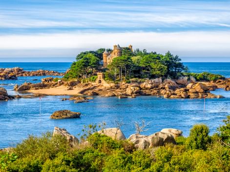 Schloss Costaérès auf einer kleinen Felseninsel, umgeben von blauem Wasser und Granitfelsen. Dichte grüne Vegetation bedeckt die Insel.