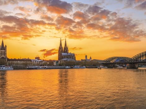 Cologne skyline at sunset with Cologne Cathedral and Hohenzollern Bridge over the Rhine. Orange and golden clouds reflect in the river water.