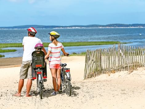 Zwei Erwachsene und ein Kind mit Fahrrädern auf einem Sandweg an der Ostsee. Sie tragen Helme und blicken auf das blaue Wasser mit grünem Marschland.