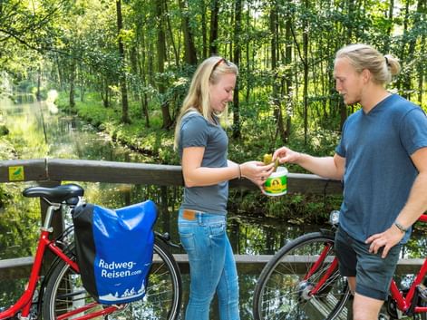 Zwei Radfahrer in blauen Shirts halten ein Glas Spreewaldgurken an einem Kanal. Rote Räder mit Radweg-Reisen-Taschen stehen neben einem Holzgeländer.