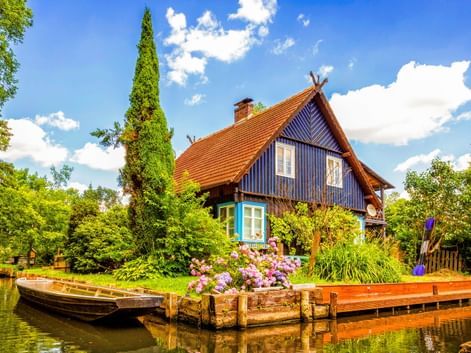 Blaues Holzhaus mit orangefarbenem Ziegeldach am Wasser im Spreewald. Holzsteg mit Blumen und Boot im Vordergrund, umgeben von üppigem Grün.