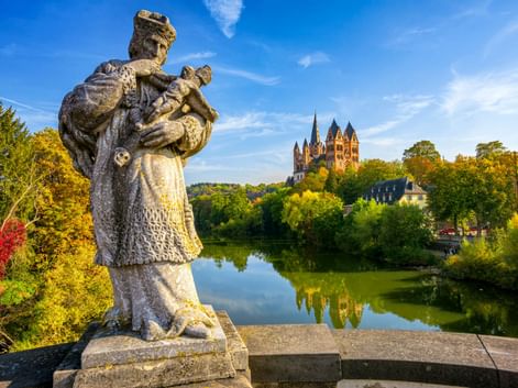 Stone statue on a bridge overlooking the Lahn River with Limburg Cathedral on a hill. Autumn trees frame the scene under a blue sky.