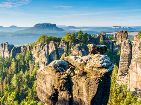 Dramatische Sandsteinfelsen der Bastei bei Rathen mit grünem Wald, fernen Tafelbergen und blauem Himmel in der Sächsischen Schweiz.