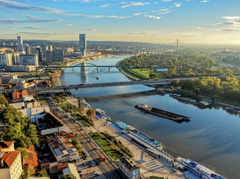 Luftaufnahme von Belgrad mit der Donau, Brücken, Kreuzfahrtschiffen und Stadtsilhouette unter blauem Himmel mit sichtbaren grünen Parks.