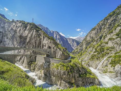 Historische Steinbrücke über die Schöllenenschlucht beim Gotthardpass. Steile Felswände rahmen einen reißenden Fluss ein, Berge im Hintergrund.