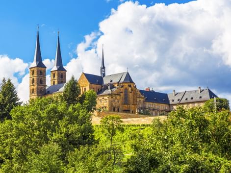 Michaelsberg Abbey in Bamberg with twin spires rising above green trees on a hilltop under a blue sky with white clouds.