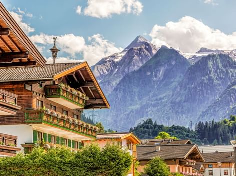 Traditional Alpine chalets in Kaprun with flower-decorated balconies, set against dramatic snow-capped mountain peaks under a blue sky with clouds.
