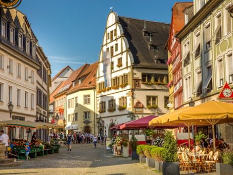 Pedestrian square in Kitzingen's old town with colorful half-timbered buildings, outdoor cafes with yellow umbrellas, and market stalls.