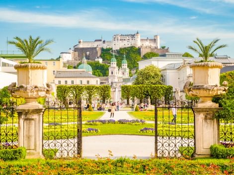 Verziertes Eingangstor mit Steinsäulen, Urnen und Palmen, das zu den Mirabellgärten mit bunten Blumenbeeten und der Festung Salzburg auf dem Hügel führt.