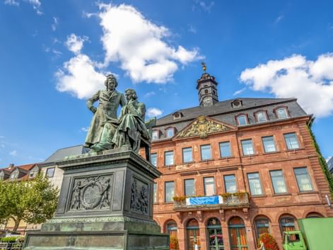 Bronzedenkmal der Brüder Grimm in Hanau mit historischem Gebäude aus rotem Sandstein dahinter. Zwei Figuren stehen auf verziertem Sockel.