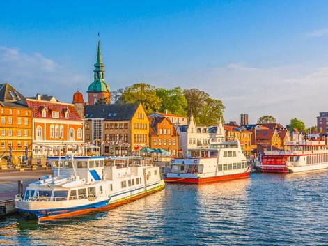 Kappeln Hafen mit weißen Passagierschiffen vor bunten historischen Gebäuden. Eine Kirche mit grünem Turm erhebt sich über orange und gelbe Fassaden.