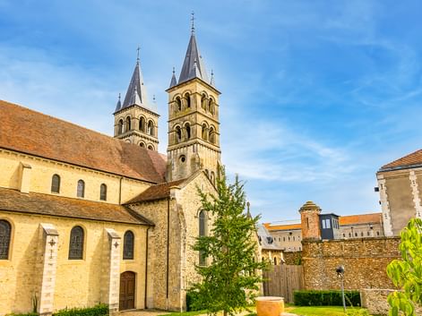 Stiftskirche Notre-Dame in Melun mit zwei romanischen Türmen mit Spitzdächern, Steinmauern und umliegenden Gebäuden unter blauem Himmel.