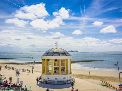 Gelb-weißer Pavillon mit Kuppel am Strand von Borkum. Sandstrand mit Besuchern, blaues Meer und bewölkter Himmel. Schiff am Horizont.