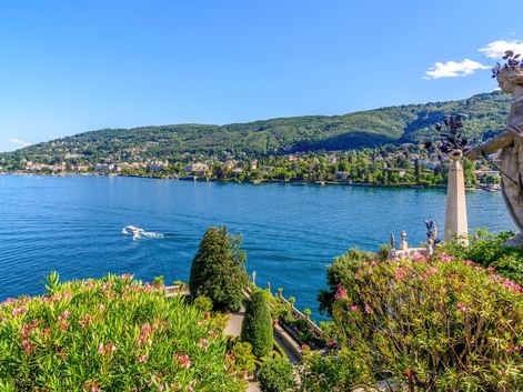 Steinstatue in einem üppigen Garten mit Blick auf das blaue Wasser des Lago Maggiore mit grünen Hügeln und einer Stadt am gegenüberliegenden Ufer.