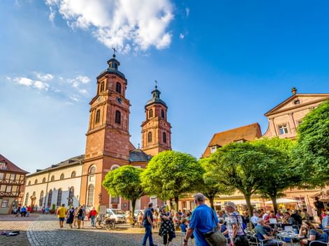 Historischer Marktplatz in Miltenberg mit Kirche mit zwei Türmen, Fachwerkhäusern und Menschen in Straßencafés unter grünen Bäumen.