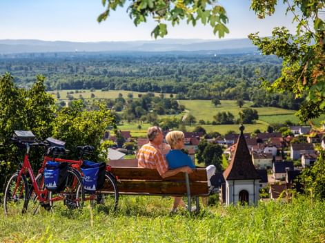 Zwei Radfahrer auf einer Bank mit Blick auf die Rheinebene bei Istein. Rote Tourenräder mit blauen Taschen stehen daneben. Dorf und grüne Felder unten.