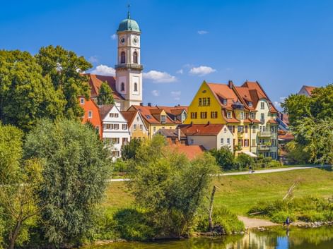 St. Mang Kirche mit weißem Turm und bunte historische Häuser in Regensburg an der Donau, umgeben von grünen Bäumen und Wiese.