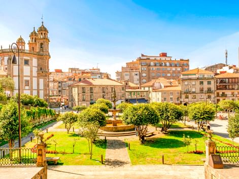 Plaza de la Herrería in Pontevedra mit grünem Park, Brunnen und historischer Kirche. Gebäude säumen den Platz unter blauem Himmel.