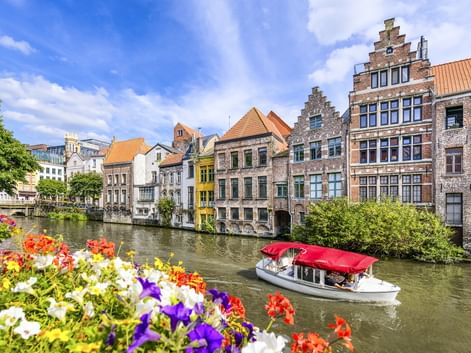 Weißes Touristenboot mit rotem Dach auf Kanal in Gent. Historische Treppengiebelhäuser säumen das Ufer, bunte Blumen im Vordergrund.
