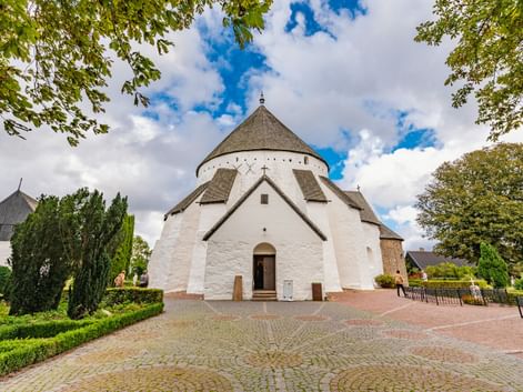 Weiße mittelalterliche Rundkirche Østerlars mit konischem Dach und Eingang, eingerahmt von grünen Bäumen unter blauem Himmel auf Bornholm.