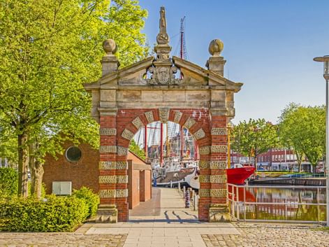 Historisches Nordertor in Emden mit rot-weißem Backsteinbogen, dekorativem Giebel und Durchblick zum Hafen mit Booten und Bäumen.