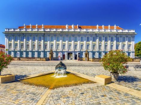 Residenz Ansbach, ein weißes Barockschloss mit orangefarbenem Dach, vom Kopfsteinpflasterplatz mit Brunnen und blühenden Pflanzen unter blauem Himmel.