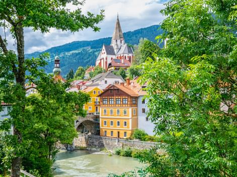 Blick auf Murau, Österreich, mit markanter Kirchturmspitze, bunten Gebäuden entlang eines Flusses und grünen Bergen im Hintergrund, eingerahmt von Bäumen.