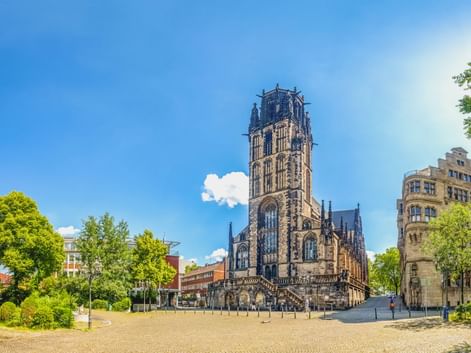 Salvatorkirche in Duisburg mit hohem gotischem Turm auf offenem Platz, umgeben von Bäumen und modernen Gebäuden unter blauem Himmel.