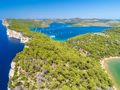 Luftaufnahme des Nationalparks Telascica mit türkisfarbener Bucht mit weißen Segelbooten, umgeben von grünen bewaldeten Klippen und felsiger Küste.