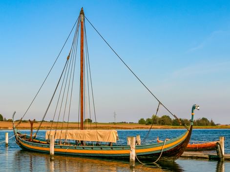 Wikingerschiff-Nachbau an einem Holzsteg auf ruhigem Wasser. Das Schiff hat einen hohen Mast, gebogenen Bug mit Drachenkopf und dunklen Rumpf mit gelber Verzierung.