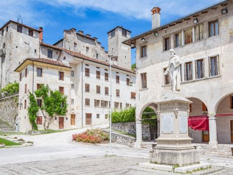 Historischer Platz in Feltre mit weißer Steinstatue von Vittorino da Feltre auf Sockel, umgeben von traditionellen italienischen Gebäuden mit Bögen.